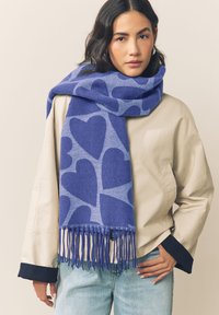 Sciarpa blu con grandi motivi a cuore in una tonalità più chiara, con bordi frangiati. Realizzata in materiale morbido, indossata sopra una giacca leggera.