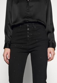 Černé džíny s vysokým pasem, se zapínáním na knoflíky a rozptýlenými čtvercovými cvočky, spárované s černou saténovou blůzou.