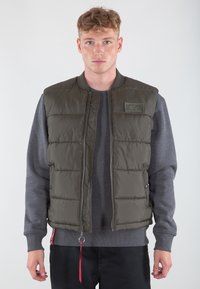 Gilet imbottito verde oliva con chiusura a zip, colletto a coste e dettaglio con toppa sul petto. Presenta un tirante rosso a contrasto e tasche laterali.