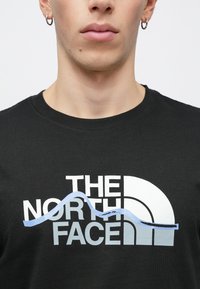 Μαύρο βαμβακερό t-shirt με το λογότυπο "THE NORTH FACE" σε λευκό και μπλε, με μινιμαλιστικό σχέδιο και απαλό ύφασμα.
