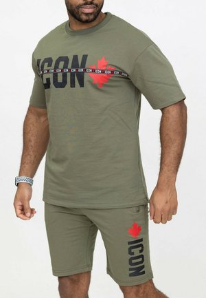 Homme portant un ensemble short et t-shirt vert olive avec une feuille d'érable rouge et le texte « ICON » sur la poitrine et la cuisse gauche.