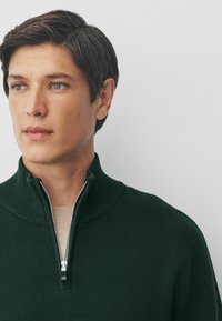 Suéter verde de punto con cuello acanalado y diseño de media cremallera; textura suave; usado sobre una camiseta beige; el rostro del modelo parcialmente visible.