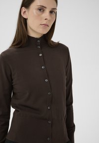 Barna kötött kardigán magas nyakkivágással és gombos elülső záródással. Bordázott textúrával és testhez simuló sziluettel rendelkezik.