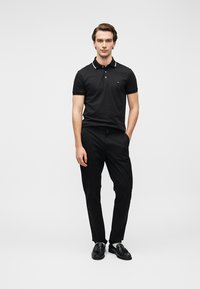 Μαύρο πόλο μπλουζάκι με λευκές λεπτομέρειες στο γιακά, συνδυασμένο με μαύρο παντελόνι και μαύρα παπούτσια. Το μοντέλο στέκεται με τα χέρια στις τσέπες μπροστά σε απλό φόντο.