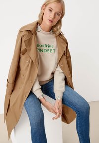 Vaaleanruskea trench-coat beigestä hupparista vihreällä tekstillä, yhdistetty sinisiin farkkuihin. Hupparissa on kuvioidut hihansuut. Yksinkertainen, moderni muotoilu.
