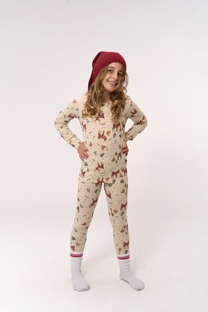Un enfant souriant se tient avec les mains sur les hanches, portant un pyjama crème avec un motif de cerf, des chaussettes blanches avec des rayures rouges et un bonnet en tricot rouge.