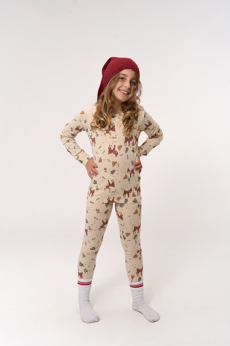 Ein lächelndes Kind steht mit auf die Hüften gestützten Händen da, trägt cremefarbene Schlafanzüge mit Reh-Muster, weiße Socken mit roten Streifen und eine rote Strickmütze.