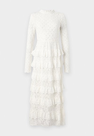 Balta mežģīņu kleita ar garām piedurknēm, augstu apkakli, daudzkārtīgu volānu apakšējo malu un ziedu rakstu visā. Tekstūras audums piešķir dziļumu.