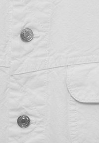 Vit plagg med texturerad tyg, silver knappar, synliga sömmar och en ficka, framhävda av geometriska sömnadsmönster.