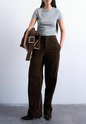 Donna che indossa un top grigio aderente a maniche corte e pantaloni larghi marrone scuro, che tiene una giacca in shearling marrone con una grande fibbia.