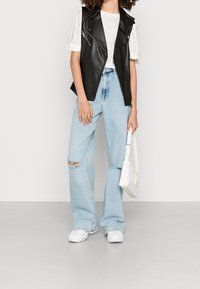 Zwart leren mouwloos vest over een witte oogjes top, gecombineerd met lichtblauwe flared jeans met gescheurde details en witte sneakers.