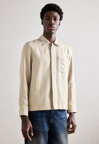 Beige vászoning gallérral, hosszú ujjakkal és egy mellzsebbel, ellazult szabással és minimalista dizájnnal, kék farmernadrággal párosítva.