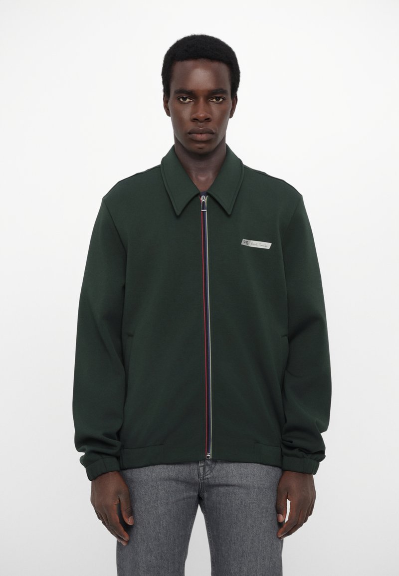 Grønn zip-up jakke med spiss krage, med et sølvmerke og fargerik stripete glidelås. Mykt stoff, avslappet passform og lange ermer.