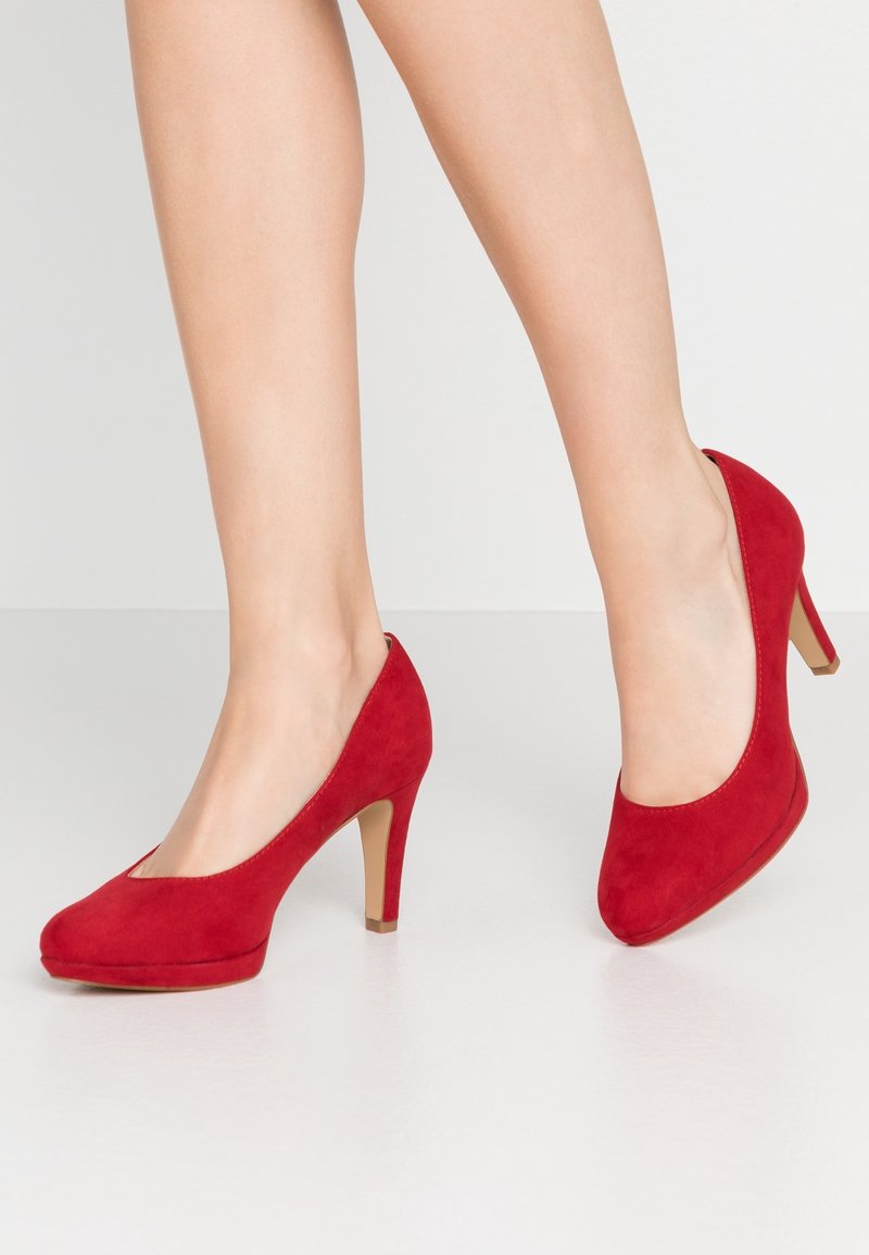 Jambes portant des escarpins hauts en daim rouge avec des bouts arrondis et des talons beiges, debout sur une surface blanche lisse.