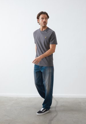 Graues Kurzarm-T-Shirt mit einem gelben Logo auf der Brust, kombiniert mit ausgebleichten blauen Jeans und schwarzen Turnschuhen mit weißen Akzenten.