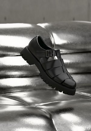 Μαύρο δερμάτινο πέδιλο με κλειστή μύτη, χοντρή σόλα και λουρί με αγκράφα, εκτεθειμένο σε γυαλιστερή ασημένια επιφάνεια με καπιτονέ υφή.