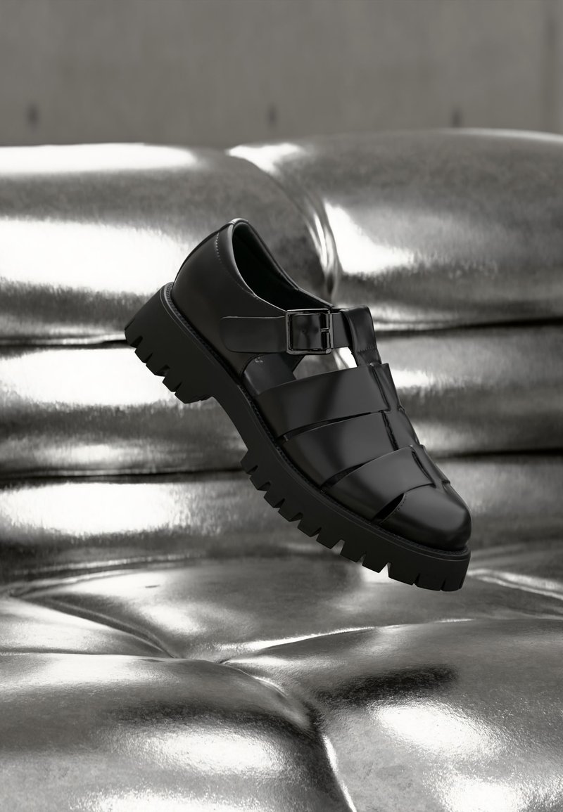 Μαύρο δερμάτινο πέδιλο με κλειστή μύτη, χοντρή σόλα και λουρί με αγκράφα, εκτεθειμένο σε γυαλιστερή ασημένια επιφάνεια με καπιτονέ υφή.