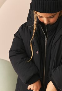 Jachetă puffer neagră cu fermoar în față și închidere cu buton; are mâneci late și umplute, cu manșete ribbate. Purtați cu o căciulă tricotată neagră.