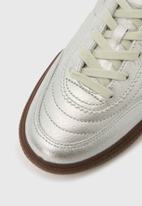 Ασημί δερμάτινη μύτη αθλητικού παπουτσιού με κρέμ κορδόνια και λεπτομερή ραφές, με καφέ υφή στην άκρη της σόλας, σε λευκό φόντο.