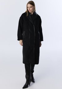 Schwarzer Mantel mit einer Kombination aus kuschliger Textur und glatten Lederpaneelen, oversized Schnitt, langen Ärmeln und zwei Vordertaschen.