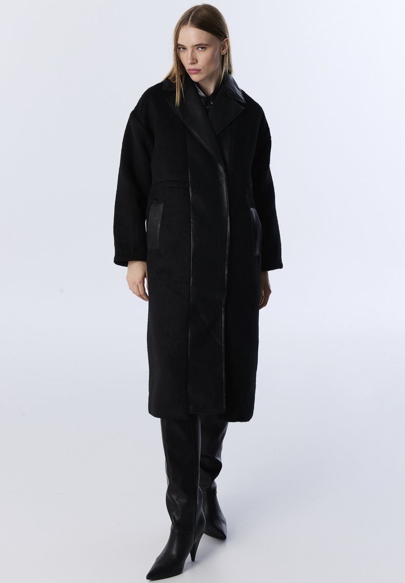 Schwarzer Mantel mit einer Kombination aus kuschliger Textur und glatten Lederpaneelen, oversized Schnitt, langen Ärmeln und zwei Vordertaschen.