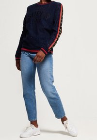 Marinblå tröja med texten "SUPER DRY", strukturerad tyg, randiga sidopaneler i röd och guld, ihopparad med ljusblå jeans och vita sneakers.