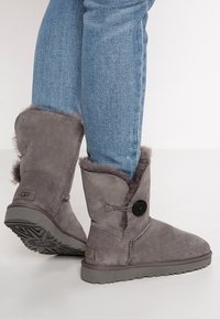 Stivaletti alla caviglia in suede grigio con fodera in pelliccia sintetica, caratterizzati da un accentuato bottone e una suola in gomma piatta. Abbinati a jeans di denim blu.