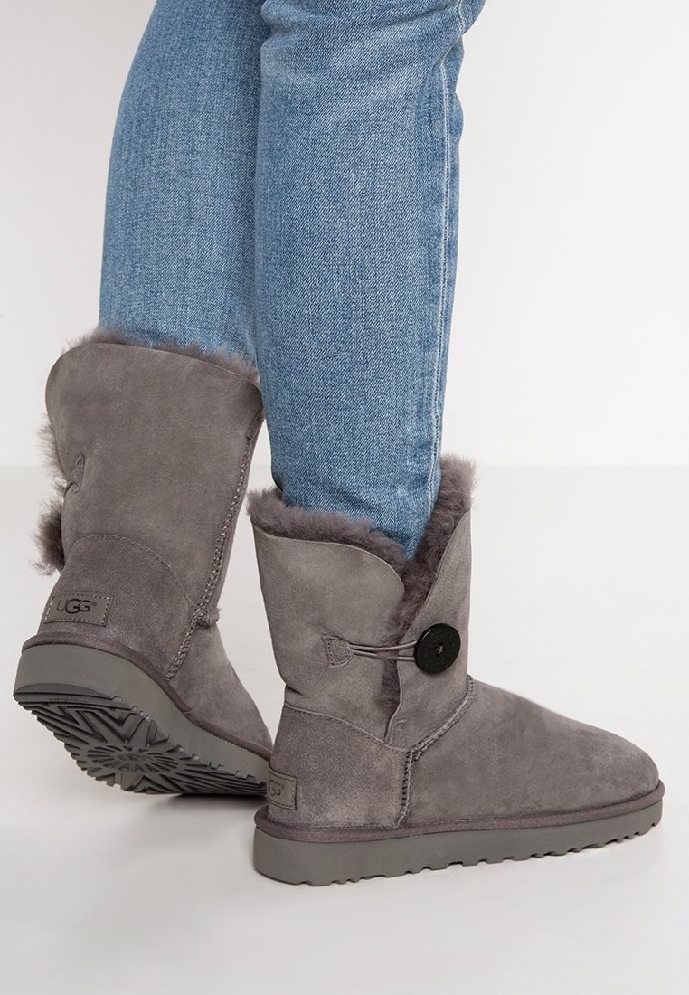 Stivaletti alla caviglia in suede grigio con fodera in pelliccia sintetica, caratterizzati da un accentuato bottone e una suola in gomma piatta. Abbinati a jeans di denim blu.