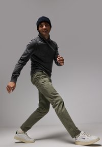 Suéter gris con cuello alto, pantalones verde oliva y zapatillas blancas con detalles en verde. Modelo en una pose dinámica de caminata.