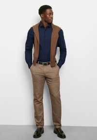 Brown sleeveless sweater draped over a navy polka-dot shirt, paired with light brown trousers and black dress shoes. Simple and tailored design.