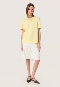 Tricou galben din bumbac cu mâneci scurte, combinat cu pantaloni scurți albi până la genunchi. Balerini negri cu vârf ascuțit și curele pentru glezne completează ținuta.