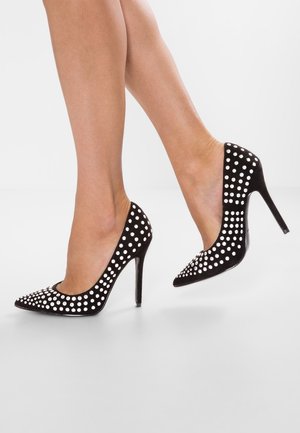 Piernas con zapatos de tacón alto negros adornados con tachuelas plateadas redondas y uniformemente espaciadas sobre un fondo blanco liso.