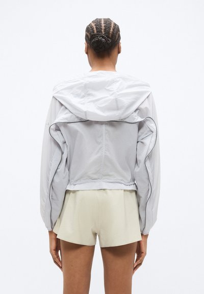 Ανοικτό γκρι μπουφάν τύπου bomber με κουκούλα, διαθέτει φαρδιά μανίκια και κοντό τελείωμα. Το ύφασμα έχει λεία, ελαφρώς γυαλιστερή υφή.