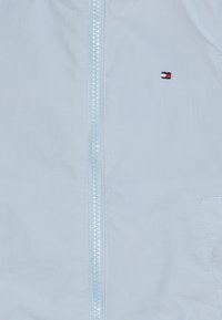 Ljusblå zip-up jacka i slätt tyg med en litenbroderad logotyp. Innehåller en central dragkedja och en sidoficka.