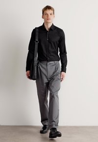 Zwarte overhemdd, grijze op maat gemaakte broek en zwarte schoenen. Het model draagt een zwarte schoudertas en staat tegen een lichte achtergrond.