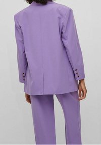 Blazer lilas ajusté en tissu lisse, avec une fente unique, manches longues ornées de boutons marron, associé à un pantalon assorti.