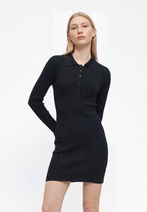 Une femme blonde porte une robe noire ajustée en maille côtelée, à manches longues, avec un col et une patte de boutonnage, sur un fond blanc uni.