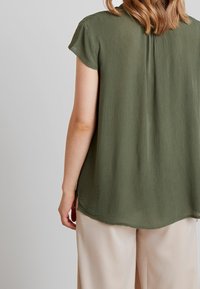 Blusa de manga corta en verde oliva con un tejido texturizado, dobladillo curvado y aberturas en los laterales. Diseñada con un sutil plisado en la parte posterior.