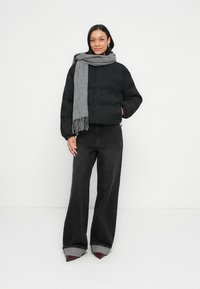 Μαύρο μπουφάν τύπου puffer με οριζόντιες ραβδώσεις, γκρι φουλάρι με κρόσσια και ψηλόμεσο σκούρο τζιν με γυρισμένα τελειώματα, συνδυασμένο με μυτερά σκοτεινά παπούτσια.