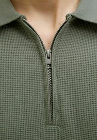 Maglietta con zip verde oliva con motivo a nido d'ape. Presenta una zip argentata e un colletto classico. Ingrandimento della scollatura e del tessuto.