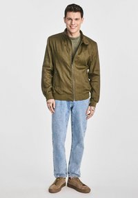 Oliwkowa bomberka z wysokim kołnierzem, zamek błyskawiczny z przodu i kieszeniami po bokach, zestawiona z jasnoniebieskimi dżinsami i brązowymi trampkami.