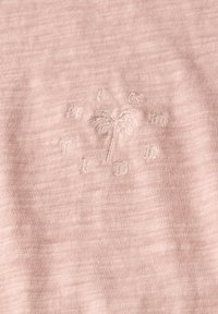 Světle růžová látka s jemnou texturou a vyšitým designem palmy a slovem "TID." Obdélníkový tvar s měkkým povrchem.