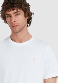 Weißes Baumwoll-T-Shirt mit rundem Ausschnitt und kurzen Ärmeln, mit einem kleinen orangefarbenen gestickten Logo auf der Brust. Glatte Textur.