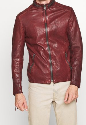 Mężczyzna ubrany w dopasowaną ciemnoczerwoną skórzaną kurtkę z zamkiem błyskawicznym i zamykanymi na zamek bocznymi kieszeniami, do tego jasnobeżowe spodnie.