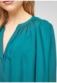Blusa teal con scollo a V profondo, spalle arricciate e morbida texture del tessuto. Presenta cuciture sottili lungo il collo e la patta.