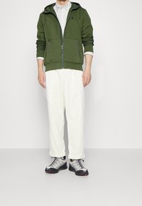 Grön zip-up hoodie, krämfärgade vida byxor och grå sneakers med röda accenter. Hoodien har sidofickor och ribbad mudd.