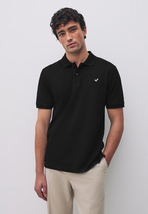 Ung mand med krøllet hår iført en sort kortærmet poloshirt med en lille fugleemblem og lyse beige bukser, stående foran en ensfarvet baggrund.