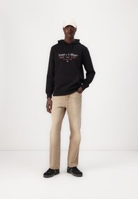 Schwarzer Hoodie mit "Tommy Hilfiger"-Logo, khakifarbene Jeans und schwarze Sneakers. Das Model trägt außerdem eine hellfarbene Mütze.