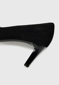 Chaussure à talon aigu en suédine noire avec un bout pointu et un talon en cuir verni brillant. Le talon mesure environ 10 cm de hauteur. Texture lisse.