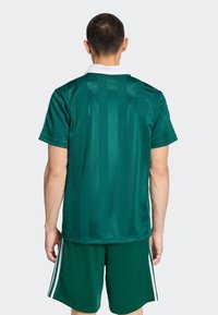 Πράσινη φανέλα με κάθετες ρίγες, κοντά μανίκια και λευκό γιακά· συνδυασμένη με πράσινα σορτς που έχουν λευκές ρίγες στο πλάι. Λείο υλικό.